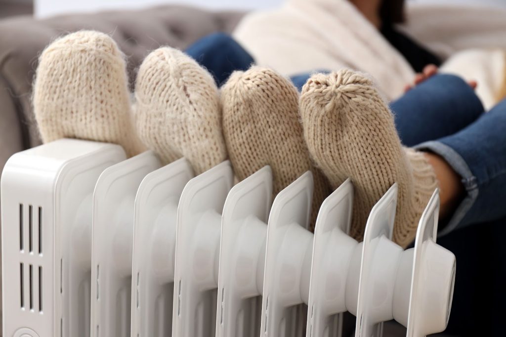 Woman in sweater and socks sitting on radiant floor heater, enjoying benefits of radiant heating
