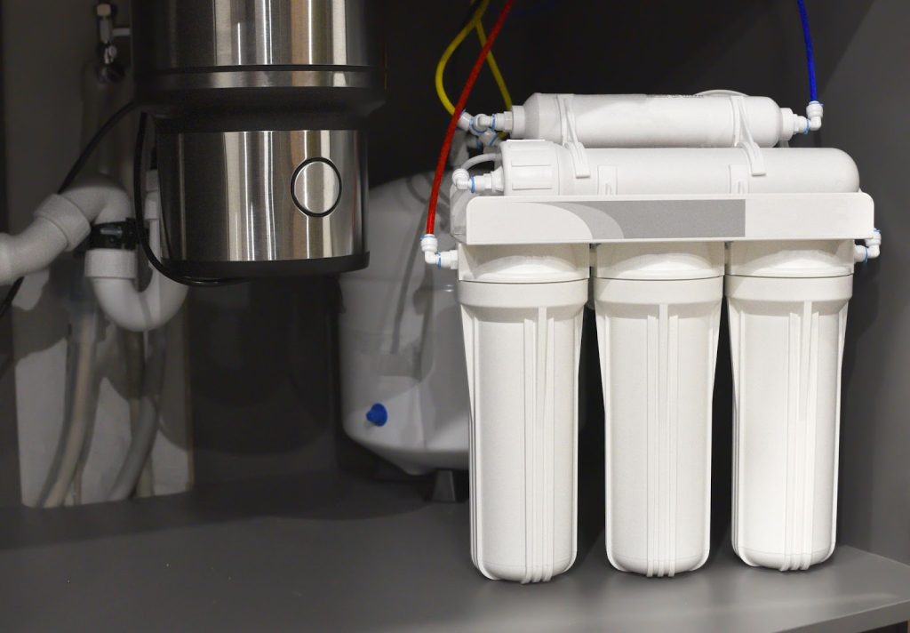 Under-sink water filtration system with multiple white filter cartridges and colored tubing.