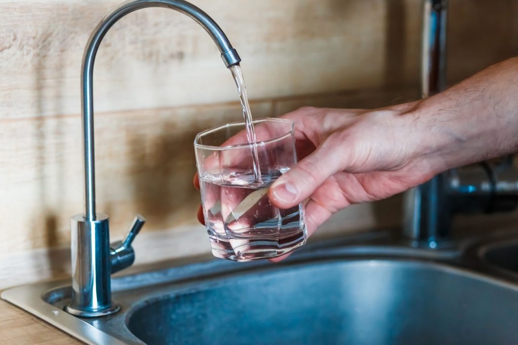 Hand filling a glass with water from a kitchen faucet over a sink.