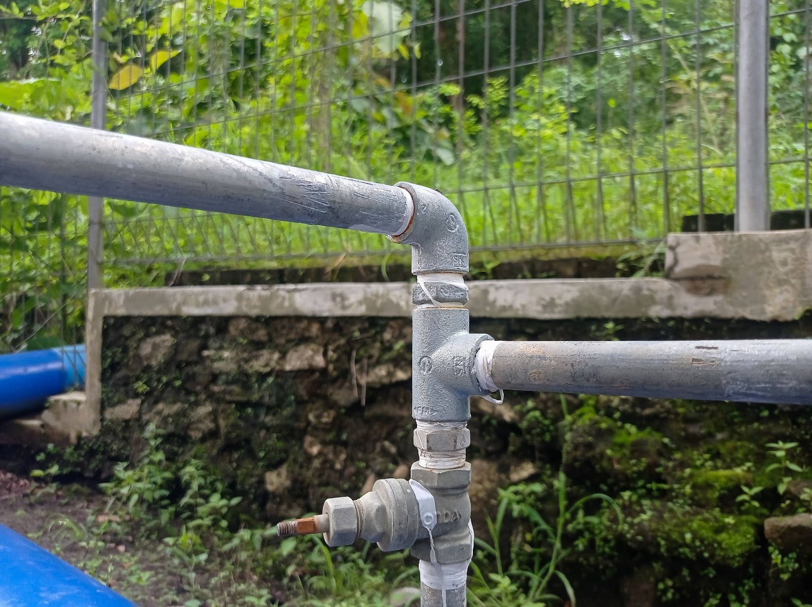 Galvanized steel pipe junction with elbow, tee, and ball valve, wrapped in white tape for sealing. Outdoor setup near concrete wall and greenery.