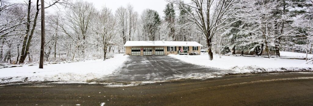 A cozy house blanketed in snow flanked by trees with a visible driveway in front