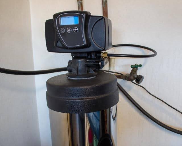 A black water softener system with a digital control panel is installed against a white wall