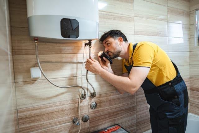 A man in a yellow shirt and blue overalls inspects a wall-mounted water heater with a flashlight