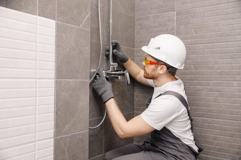 A man wearing a hard hat and safety glasses repairs a shower head in a bathroom setting