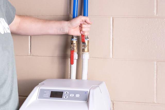 A person in a green shirt adjusts red valves on blue and white pipes connected to a device set against a beige brick wall