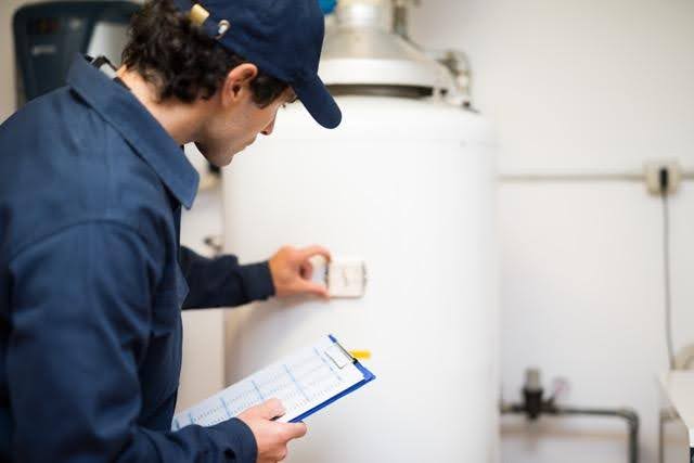 A technician in a blue uniform inspects a white water heater checking controls while holding a clipboard