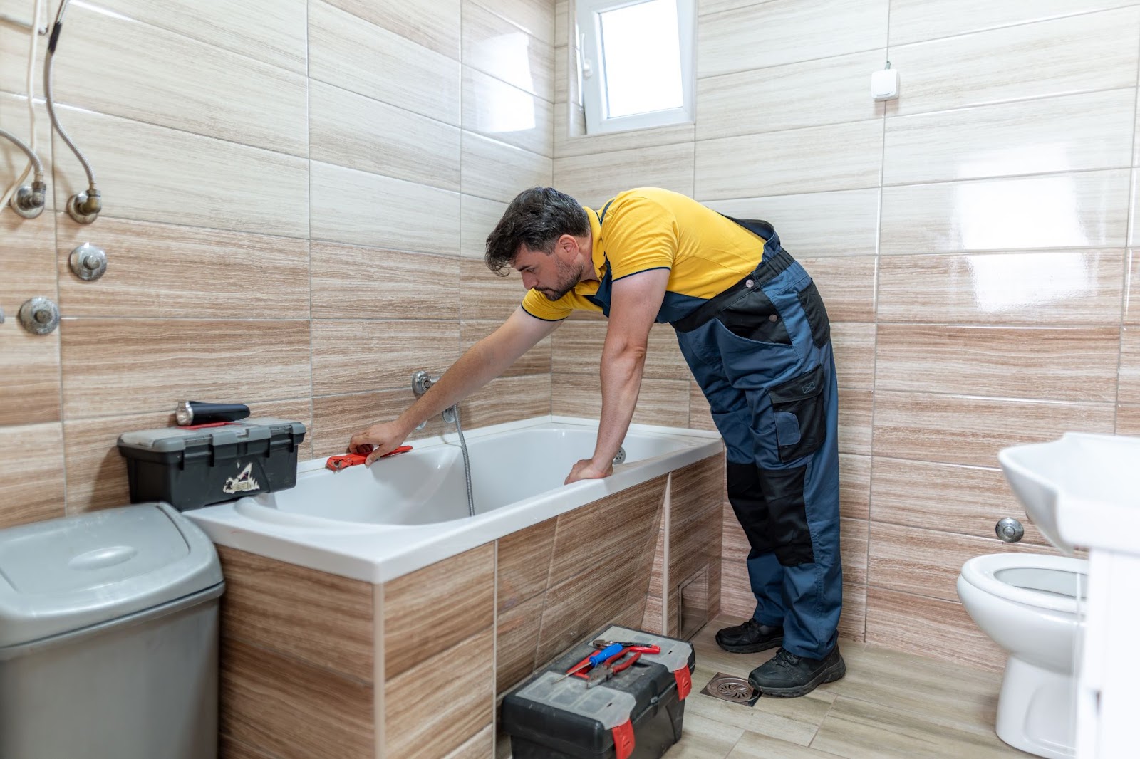 A plumber in a yellow shirt and blue overalls uses a pipe wrench to work on a bathtub fixture.