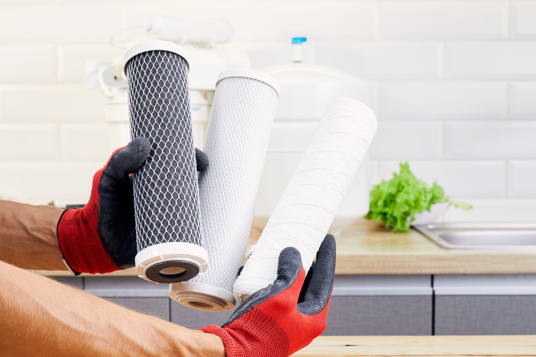 Gloved hands holding three different replacement water filter cartridges in a kitchen.